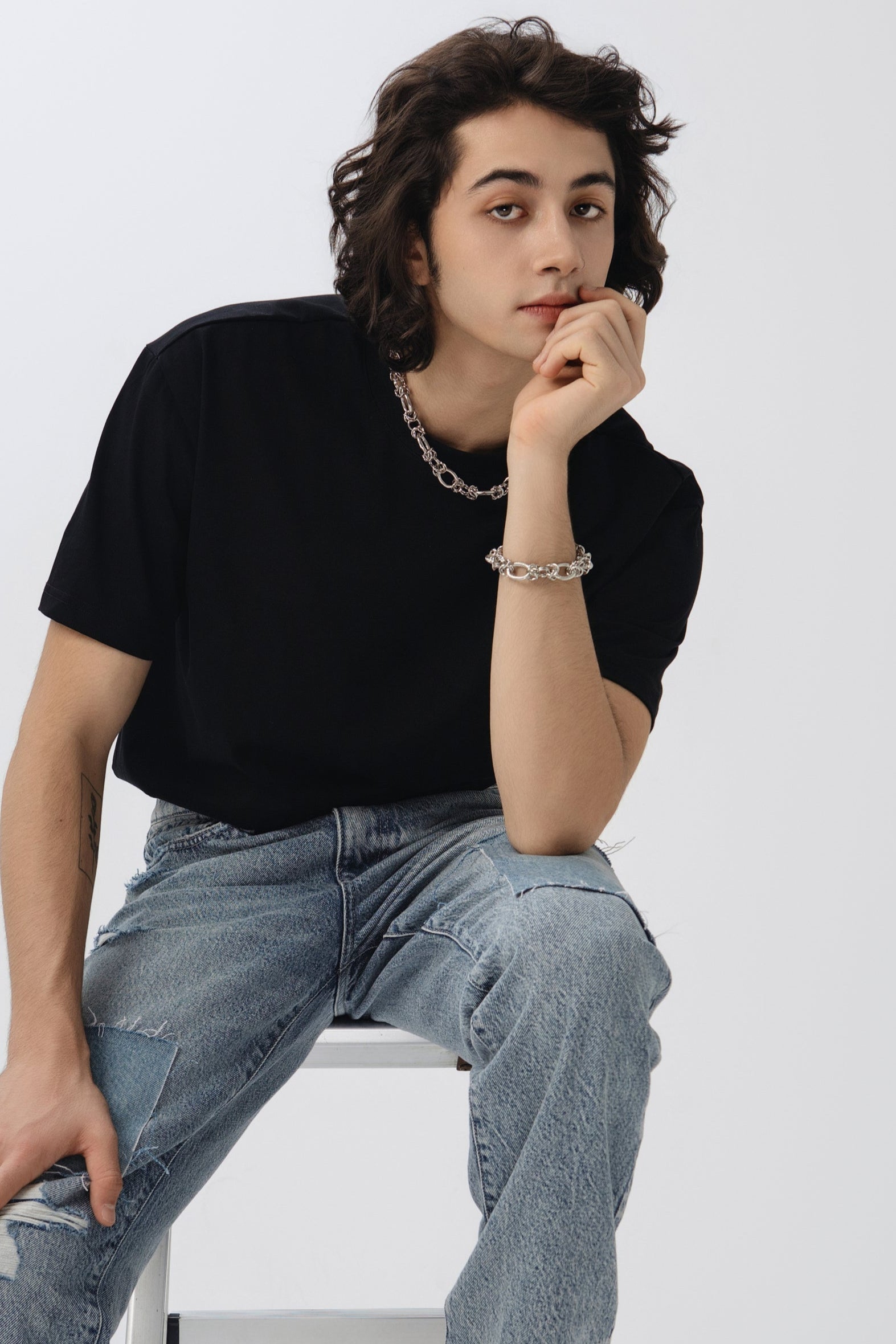 Person wearing a black t-shirt and blue jeans sitting on a white stool against a white background.